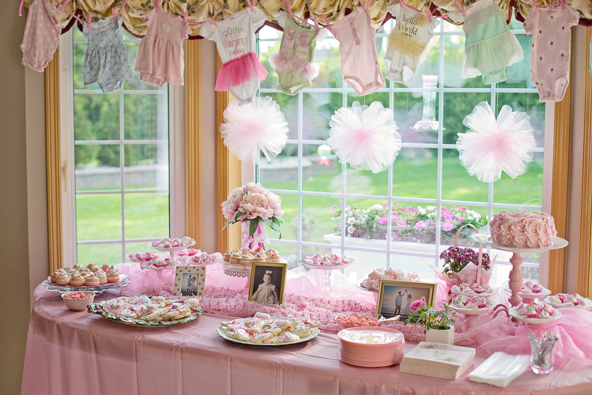 pink themed food table backlight by a big window that has puff balls hanging in it and cute onesies haging above it | 12 Steps to Hosting the Perfect Baby Shower by Busy Life Healthy Wife